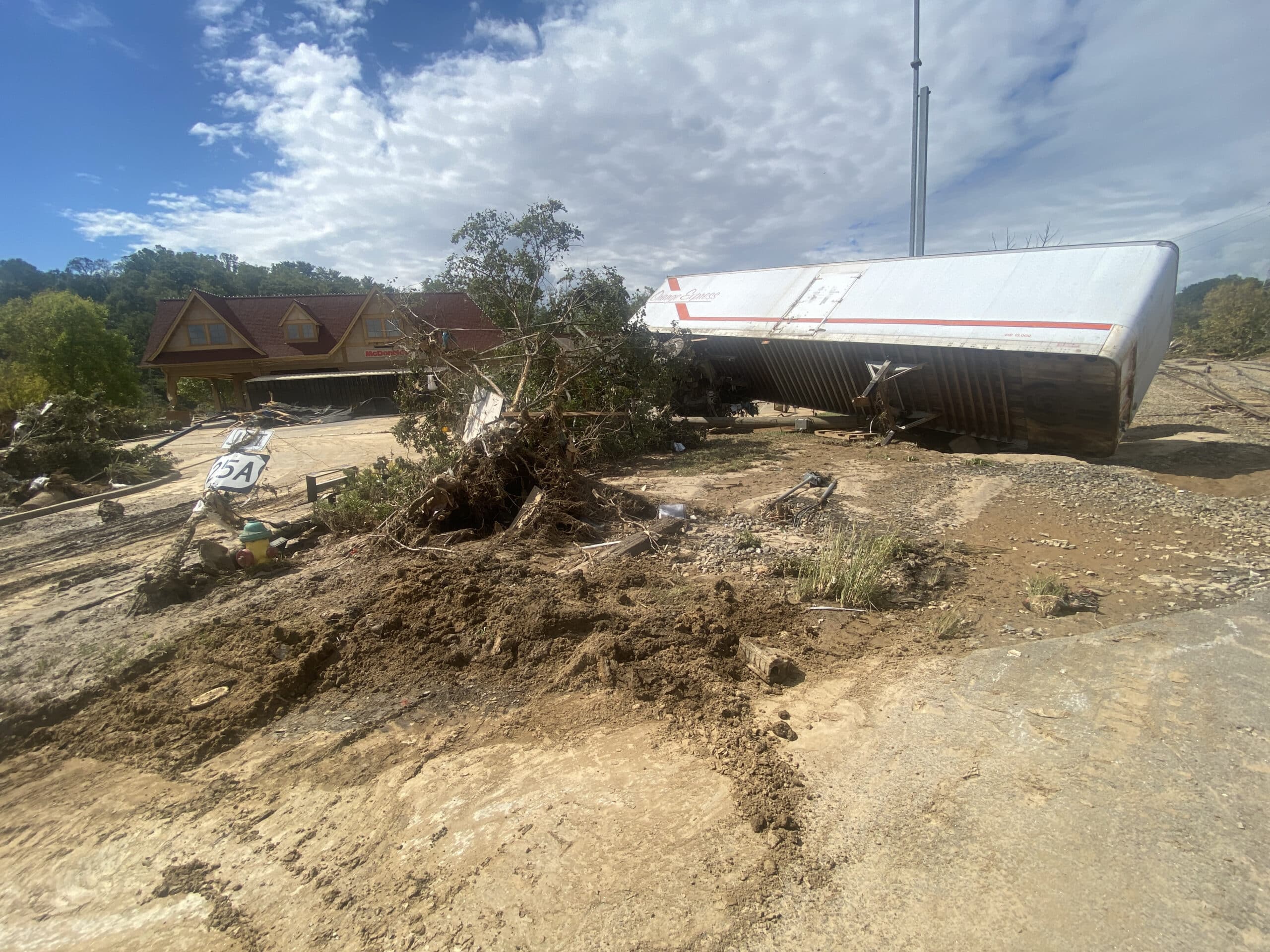 A large overturned trailer lies on a dirt path, surrounded by debris and uprooted trees. In the background, a house with a steep roof stands under a partly cloudy sky. Signs of flooding are visible on the ground.