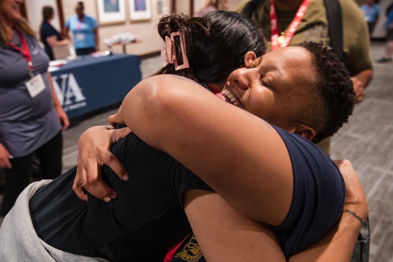 Two people warmly embrace each other in a room with a table covered by a blue cloth in the background. Both appear joyful, suggesting a reunion or celebration. Other people can be seen standing behind them.