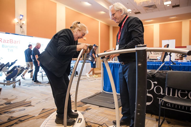 A woman standing on a vibrating exercise platform smiles while a man in a suit discusses the device with her. They are in a convention hall with exhibitors and attendees in the background.