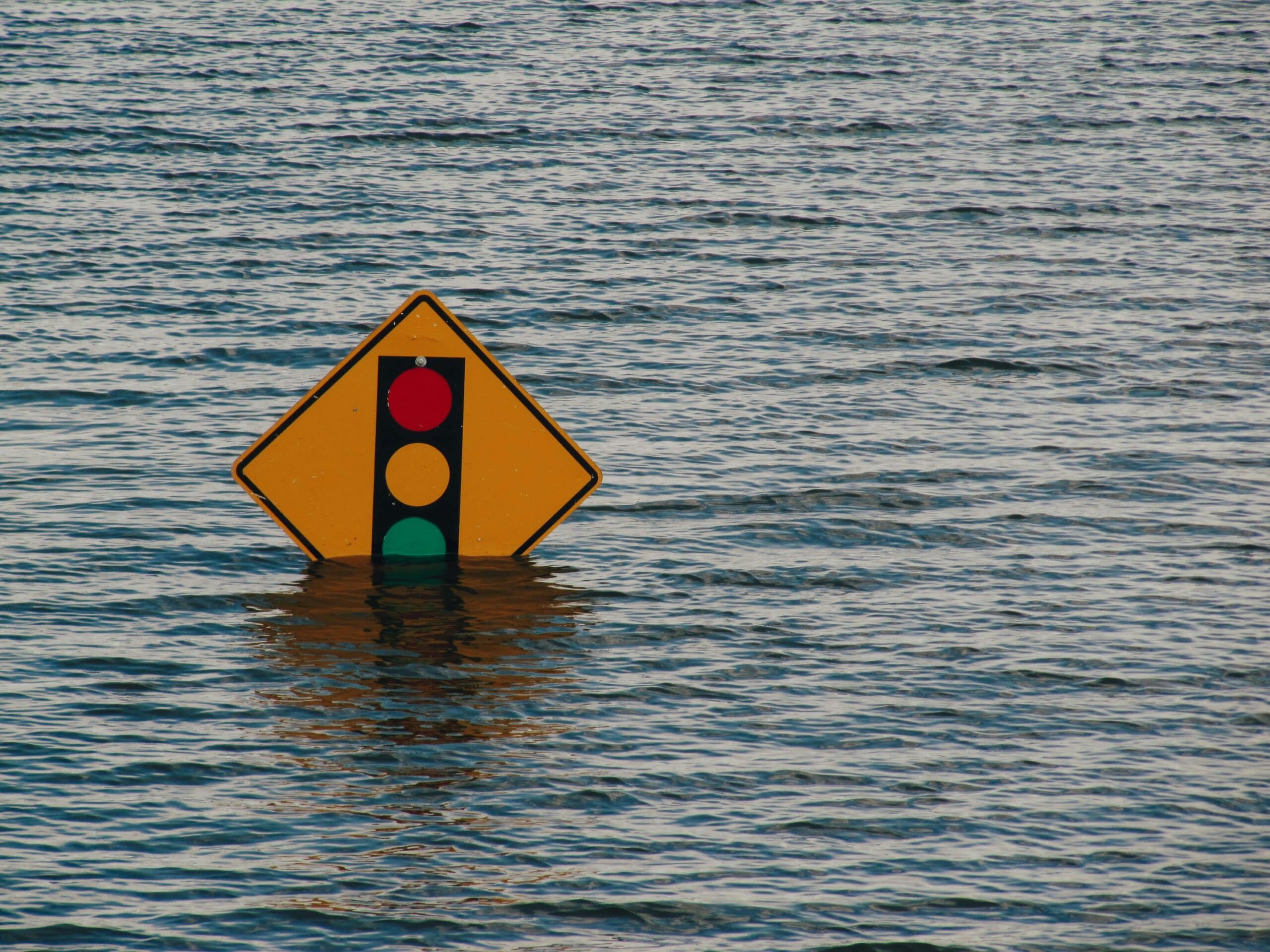 A yellow traffic sign depicting a traffic light is partially submerged in water, surrounded by rippling waves, suggesting flooding.