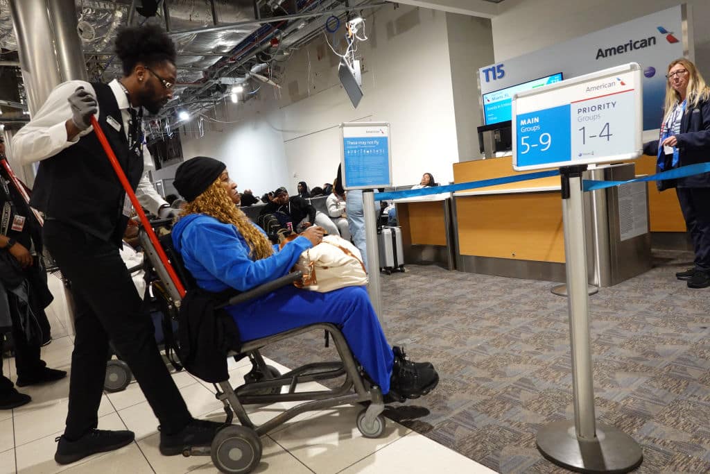 A man assists a woman in a wheelchair at an airport terminal near boarding gates. Travelers and airline staff are visible in the background. Signs indicate main and priority boarding groups.