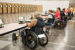 Athletes in wheelchairs participate in a shooting event, aiming at targets in an indoor range. They are seated at tables, each with a pistol and a monitor displaying their aim. The background shows a series of target boards.