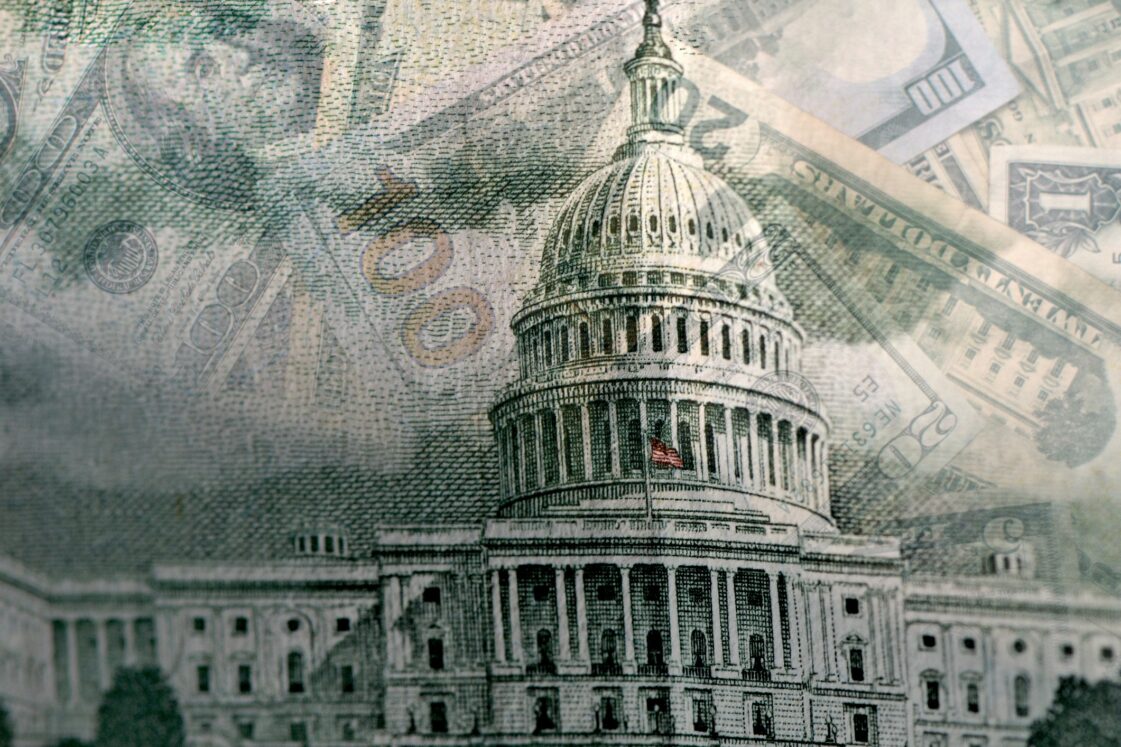 Close-up of the U.S. Capitol building with superimposed images of various denominations of U.S. currency, including 100-dollar bills. The composition suggests themes of government finance and economics.