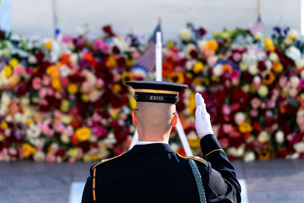 A member of the military in a ceremonial uniform salutes in front of a vibrant floral display with American flags. The scene conveys a solemn and respectful tribute.