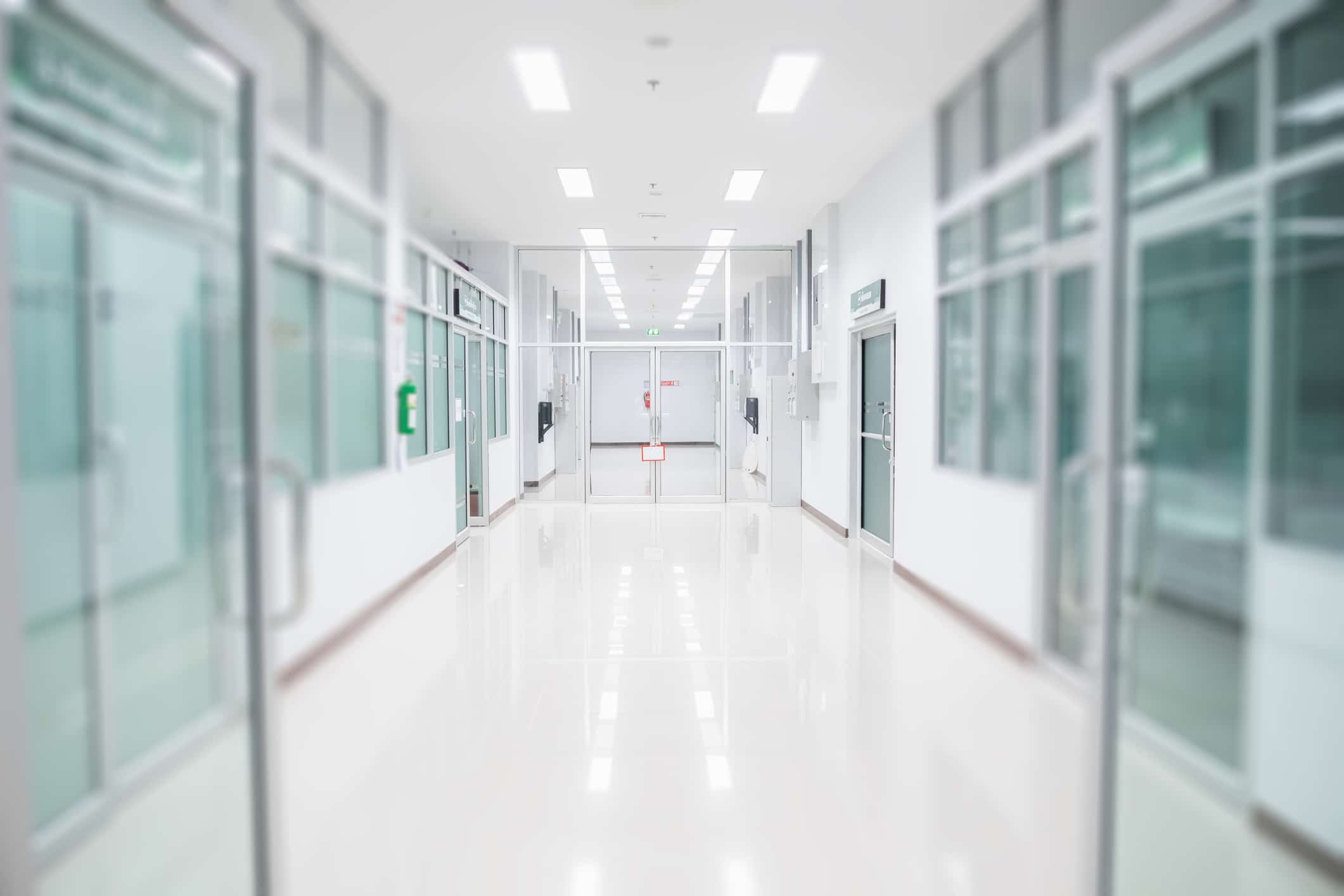 A clean, bright hospital corridor with shiny white floors and fluorescent lighting. Glass doors line both sides, and a red fire extinguisher is visible on the left wall. The space feels sterile and modern.