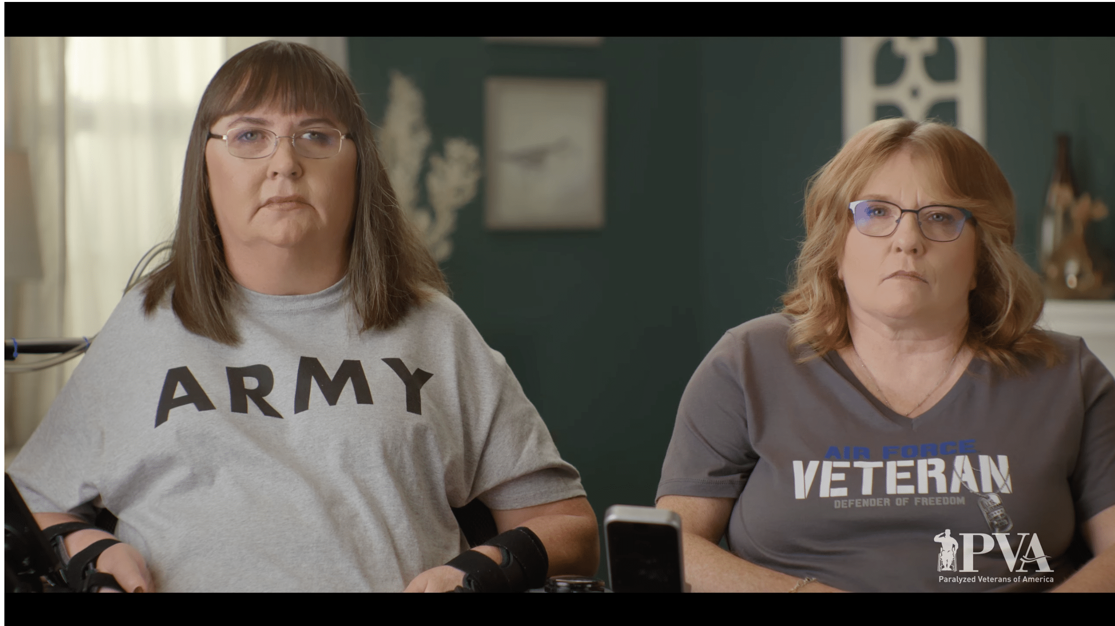 Two women sit side by side. One wears a gray "ARMY" T-shirt and uses a wheelchair, while the other wears a gray "AIR FORCE VETERAN" T-shirt. Both have serious expressions. The PVA logo is visible in the corner.