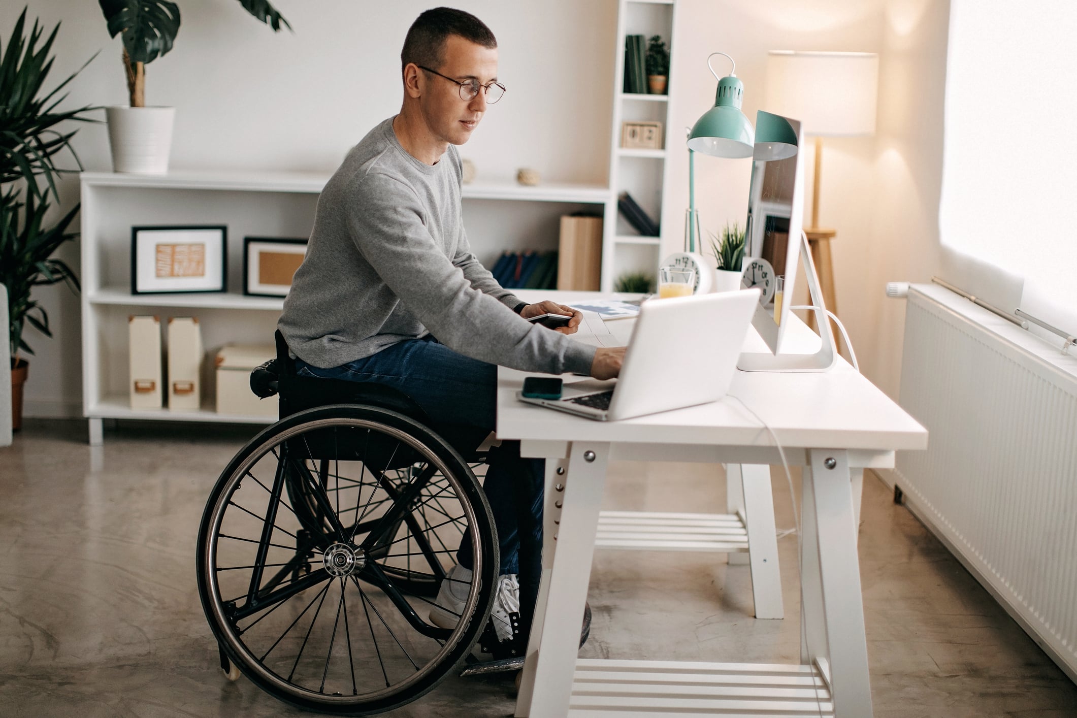 A man using a wheelchair is working at a desk with a computer and a laptop. The room has a modern design with shelves, plants, and framed pictures. Natural light illuminates the workspace from a nearby window.
