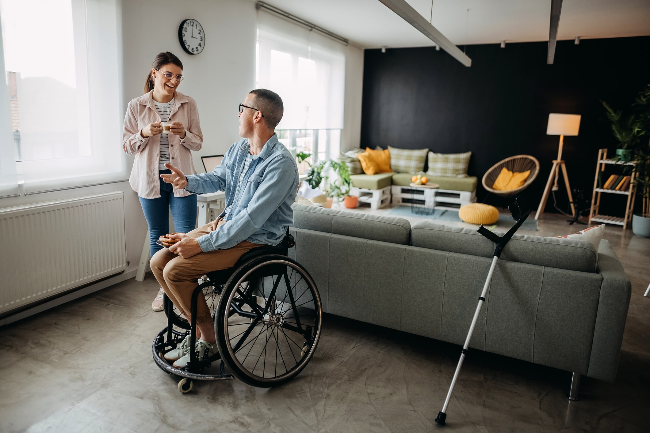 A man in a wheelchair holds a phone and talks to a woman standing beside him, both smiling in a modern living room. A crutch leans against the sofa, and a clock, plants, and a lamp are in the background.