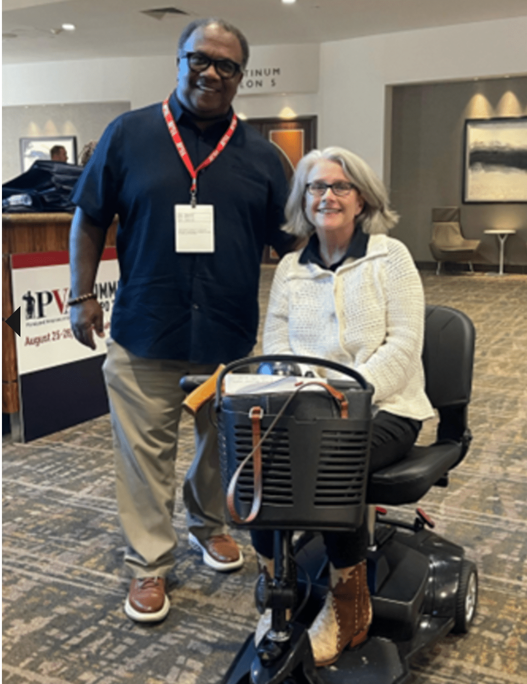 A man in a blue shirt and a woman with gray hair and glasses, wearing a light-colored sweater, sit on a mobility scooter indoors. They both smile at the camera, standing on a carpeted floor near a conference sign.