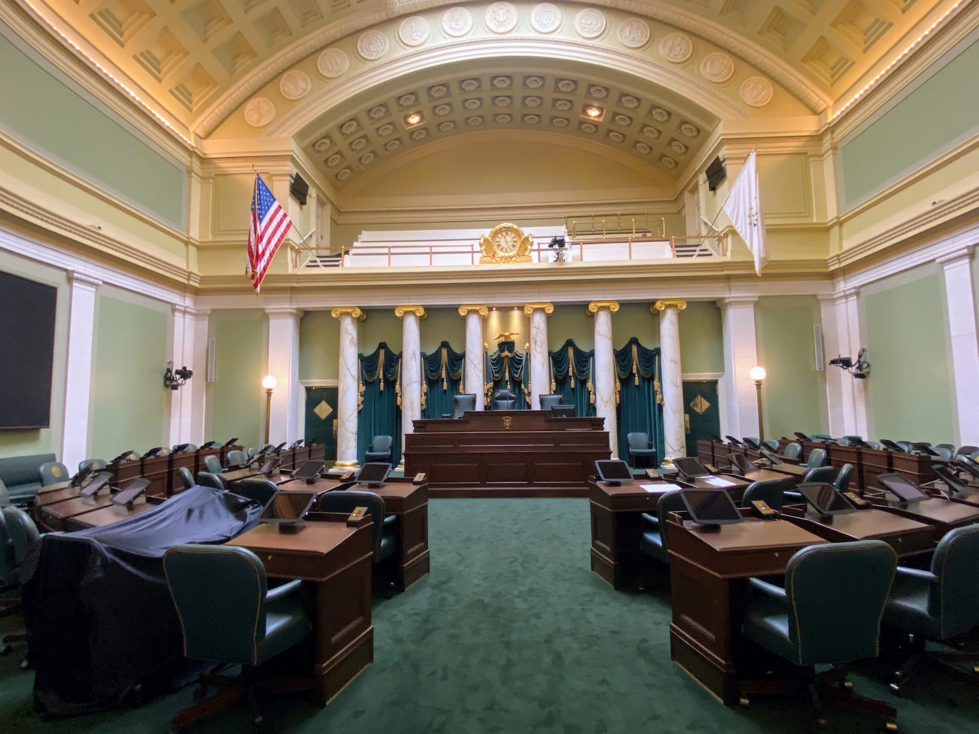 A legislative chamber with green carpeting, featuring dark wood desks and green chairs. The room has a large, decorated ceiling, an American flag, and a central elevated area with a podium and curtains.