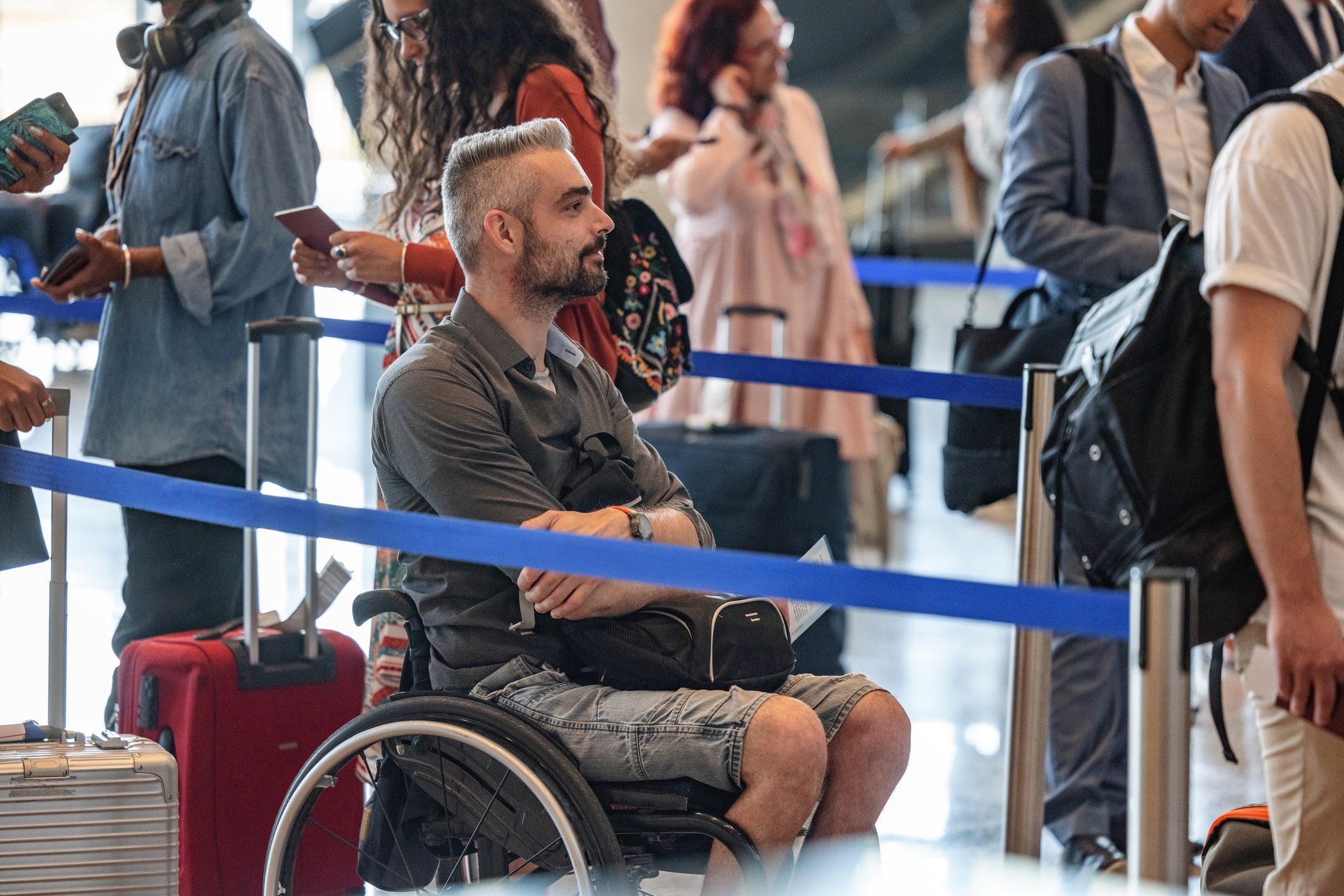 A man in a wheelchair, wearing a gray shirt and denim shorts, waits in an airport line with other travelers. He has a silver suitcase and a backpack. People are engaged with their devices and luggage around him. Blue ropes guide the queue.