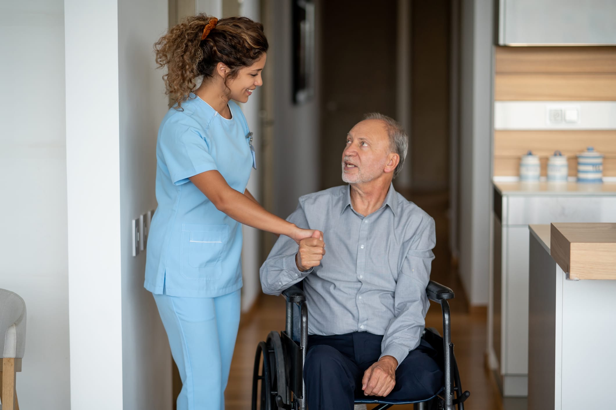 A caregiver in a blue uniform assists an elderly man in a wheelchair. They are in a well-lit room with a kitchen in the background. The caregiver is smiling while holding the man's hand, and he looks up at her appreciatively.