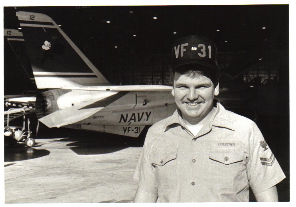 George Holscher, U.S. Navy Veteran, in military uniform, smiles in front of a jet inside a hangar. The jet's tail has "Navy" and "VF-31" markings. George wears a cap also labeled "VF-31.