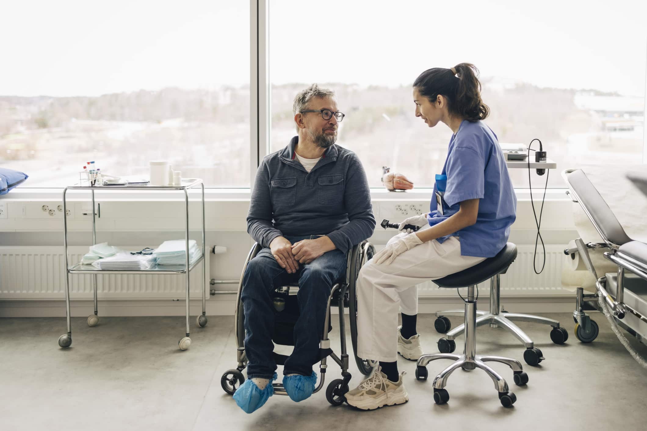 A caregiver in a blue uniform assists an elderly man in a wheelchair. They are in a well-lit room with a kitchen in the background. The caregiver is smiling while holding the man's hand, and he looks up at her appreciatively.