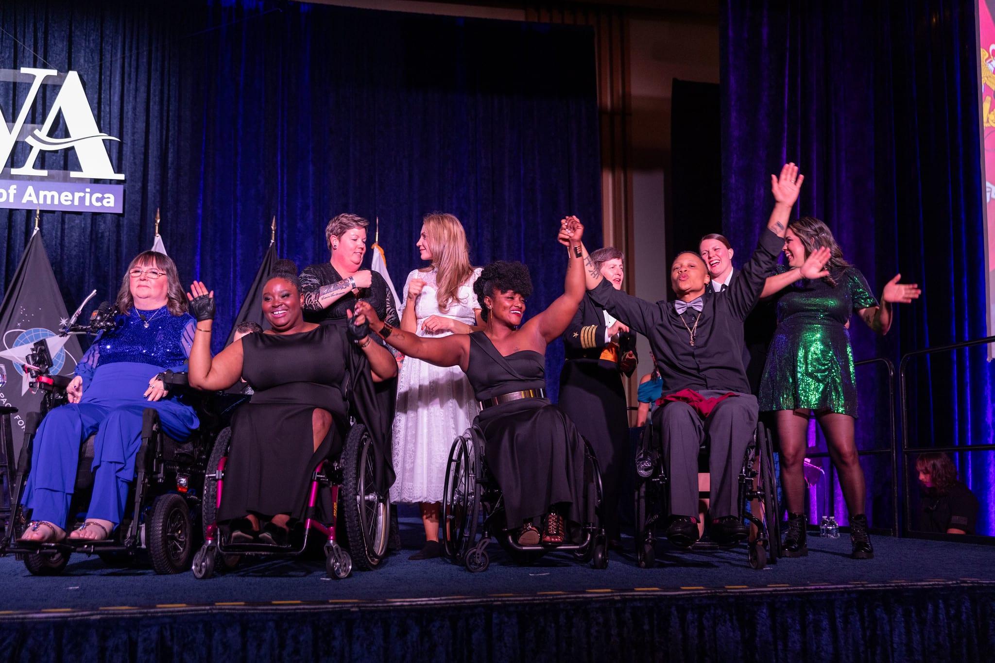 A group of smiling people on stage, some in wheelchairs, holding hands triumphantly. They are dressed in formal attire, with a dark blue curtain as a backdrop. A sign reading "WA" is partially visible on the left.