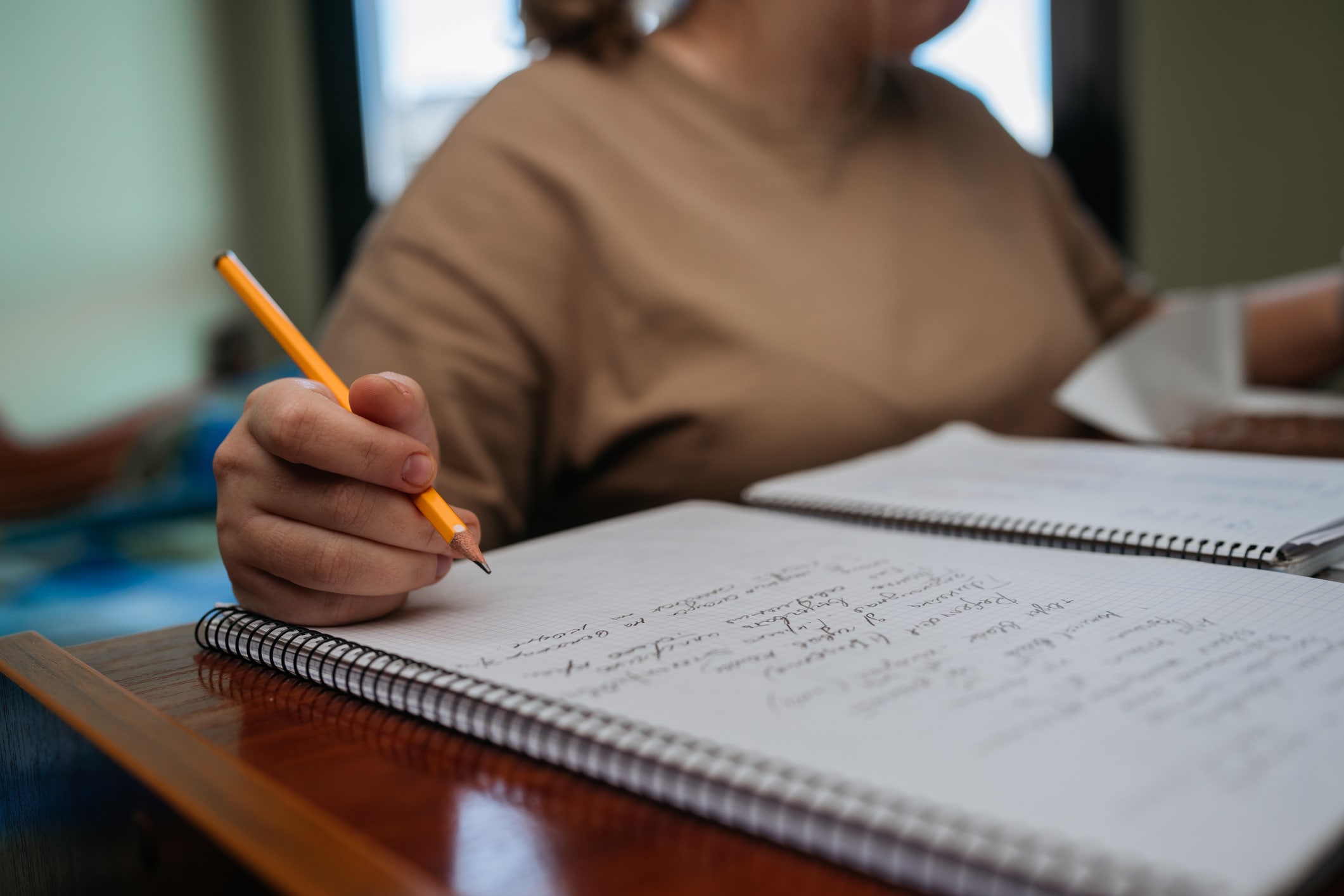 A man in a wheelchair, wearing a gray shirt and denim shorts, waits in an airport line with other travelers. He has a silver suitcase and a backpack. People are engaged with their devices and luggage around him. Blue ropes guide the queue.