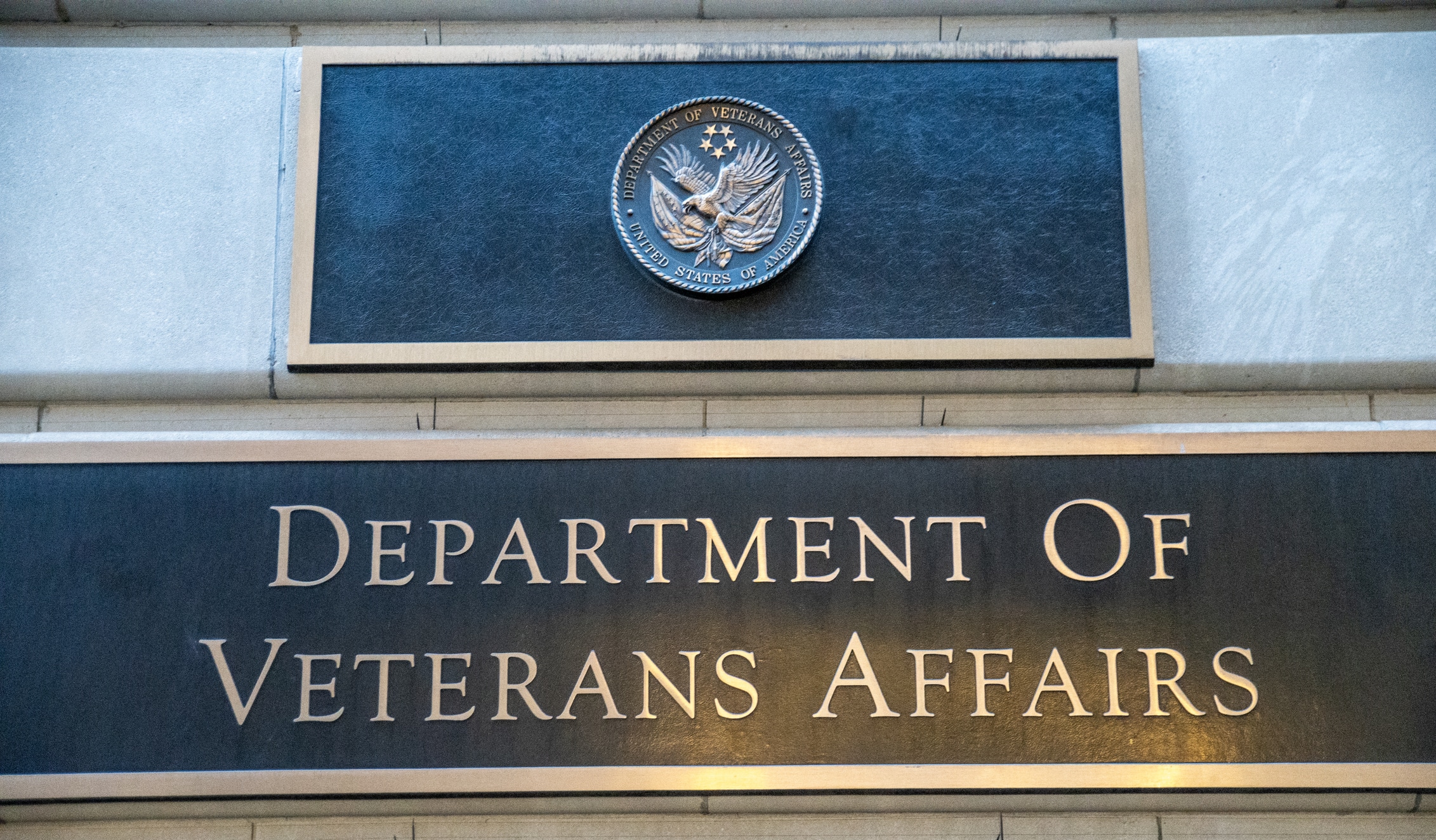 A man in a wheelchair, wearing a gray shirt and denim shorts, waits in an airport line with other travelers. He has a silver suitcase and a backpack. People are engaged with their devices and luggage around him. Blue ropes guide the queue.