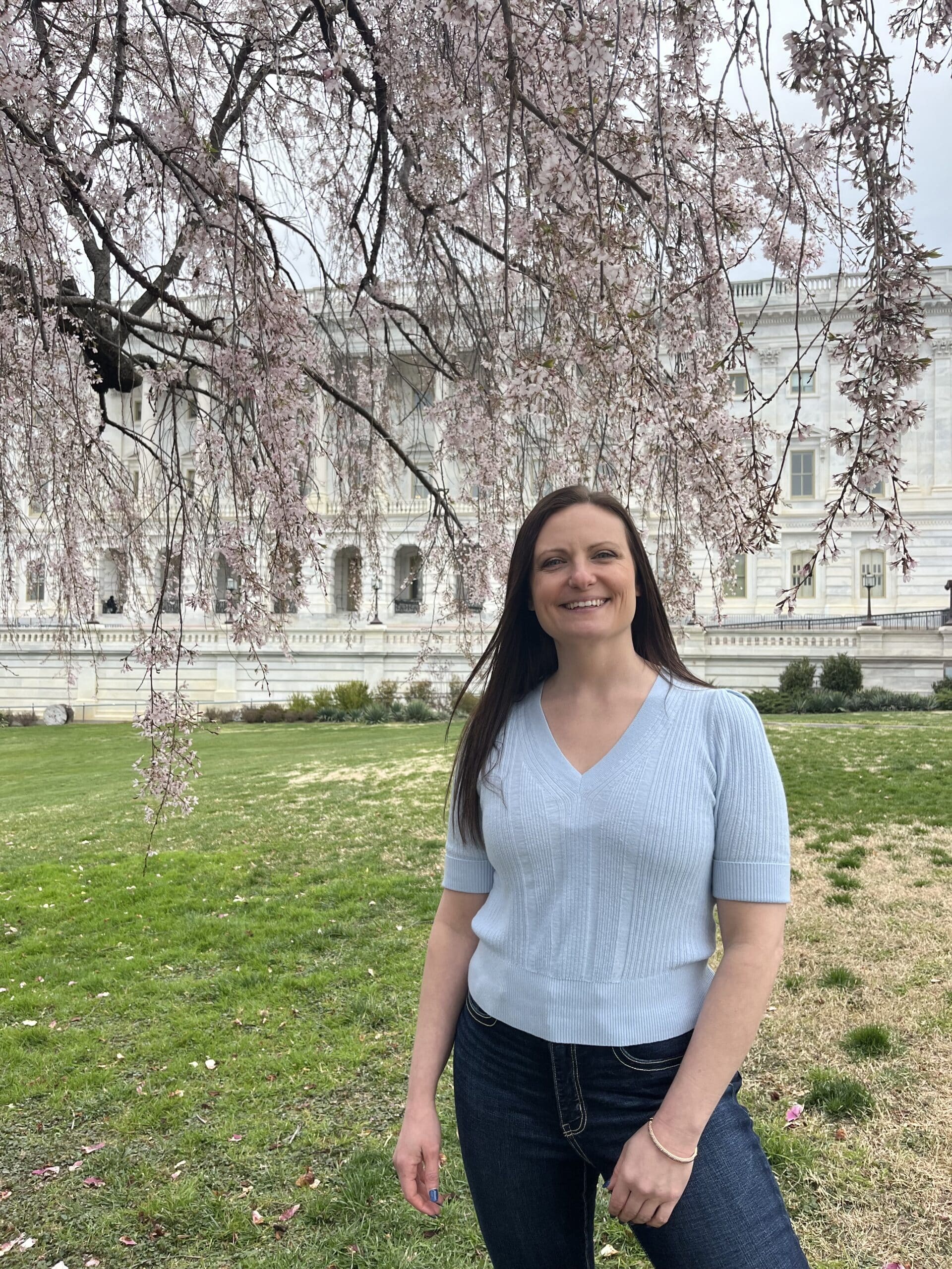 A woman stands smiling on a grassy area with tree branches above. She wears a light blue top and jeans, with a white building in the background.