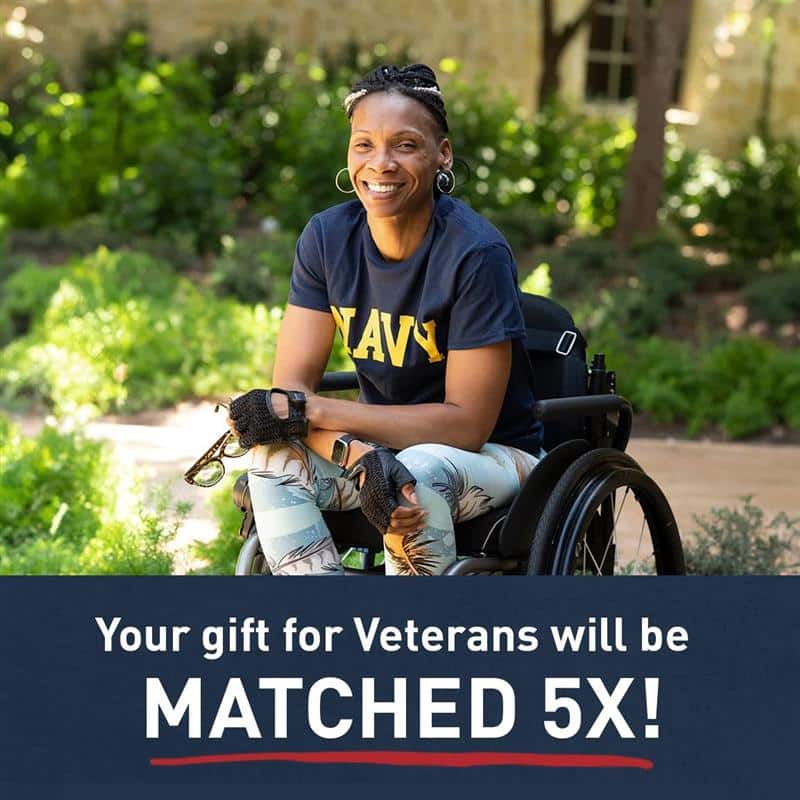 A woman stands smiling on a grassy area with tree branches above. She wears a light blue top and jeans, with a white building in the background.