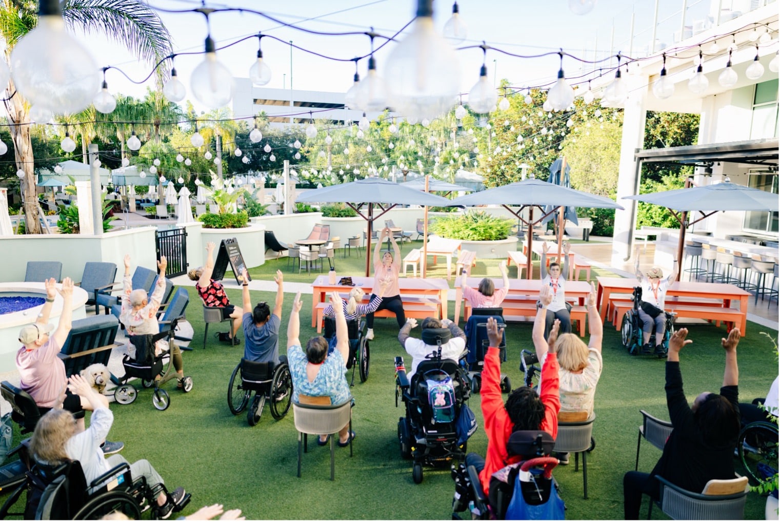 A group of seniors, some in wheelchairs, participate in an outdoor exercise class, raising their arms under string lights on a sunny patio surrounded by greenery.