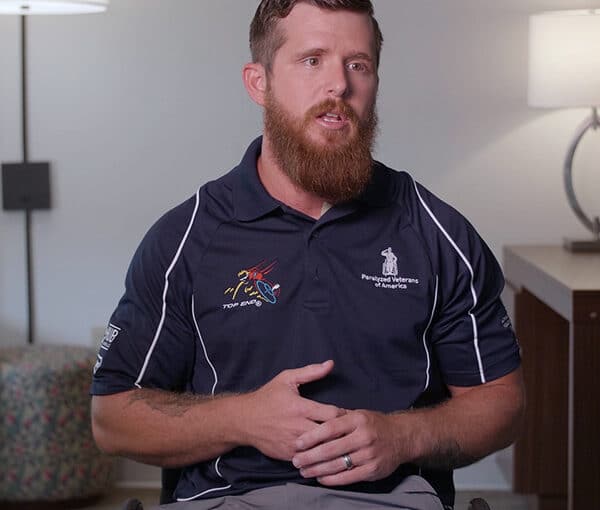 A man with a beard sits indoors, wearing a navy Paralyzed Veterans of America polo shirt. He is speaking and gesturing with his hands, and a lamp and furniture are visible in the background.