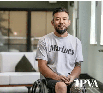 A man in a wheelchair wearing a gray "Marines" t-shirt sits and smiles indoors. The Paralyzed Veterans of America (PVA) logo is in the lower right corner.