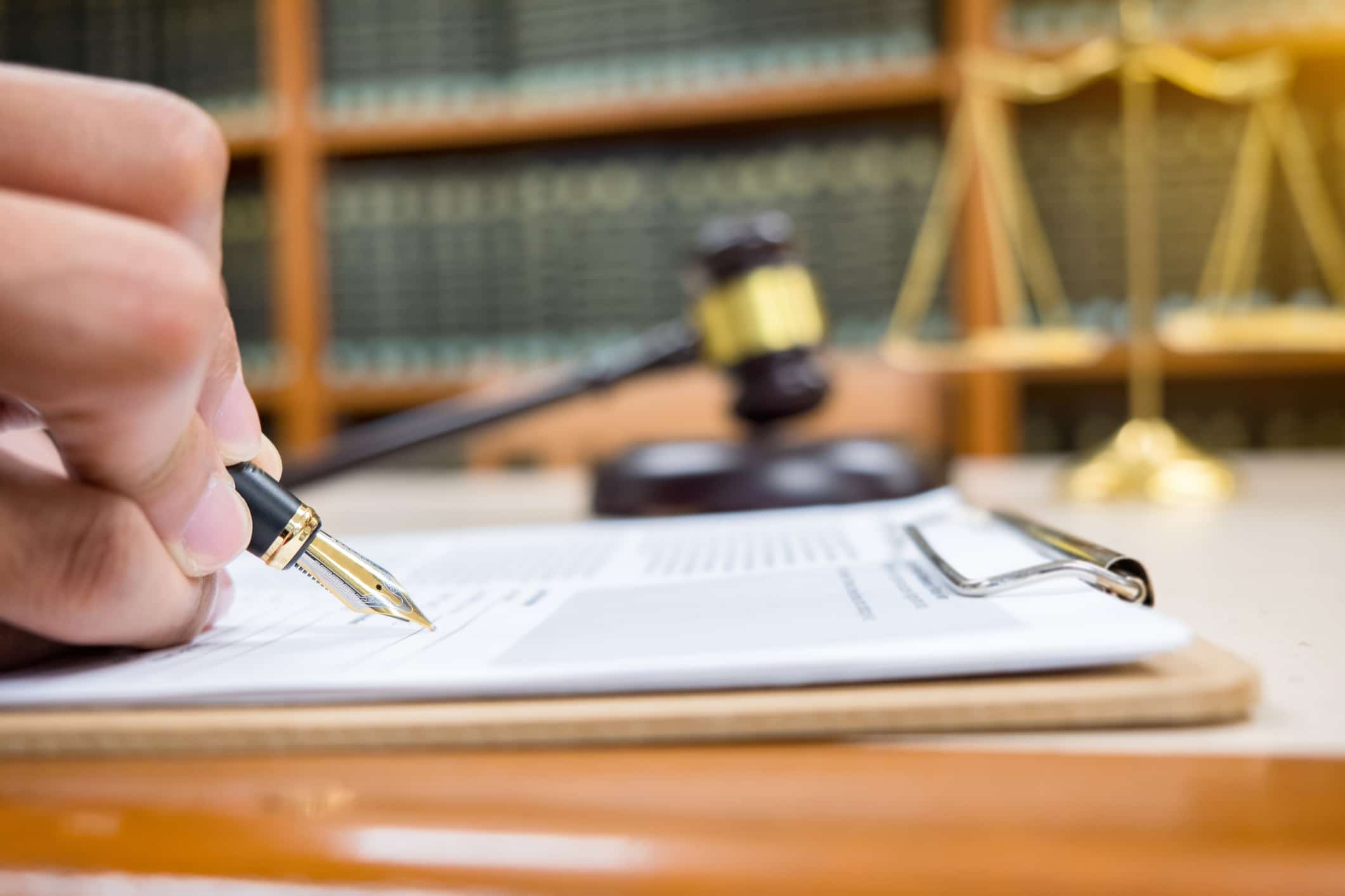 A hand holding a pen signs a document on a clipboard, with a judge’s gavel, scales of justice, and law books visible in the background, suggesting a legal or courtroom setting.