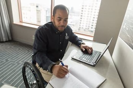 A man in a wheelchair is sitting at a desk, using a laptop and writing in a notebook, with a cityscape visible through the window behind him.
