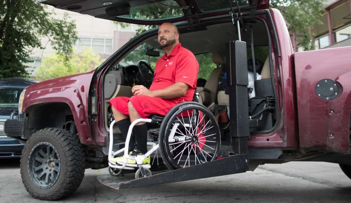 A man in a wheelchair uses a lift to exit a red pickup truck. He is wearing a red shirt and shorts, and the truck door is open, showing an accessible vehicle modification. Trees and buildings are in the background.