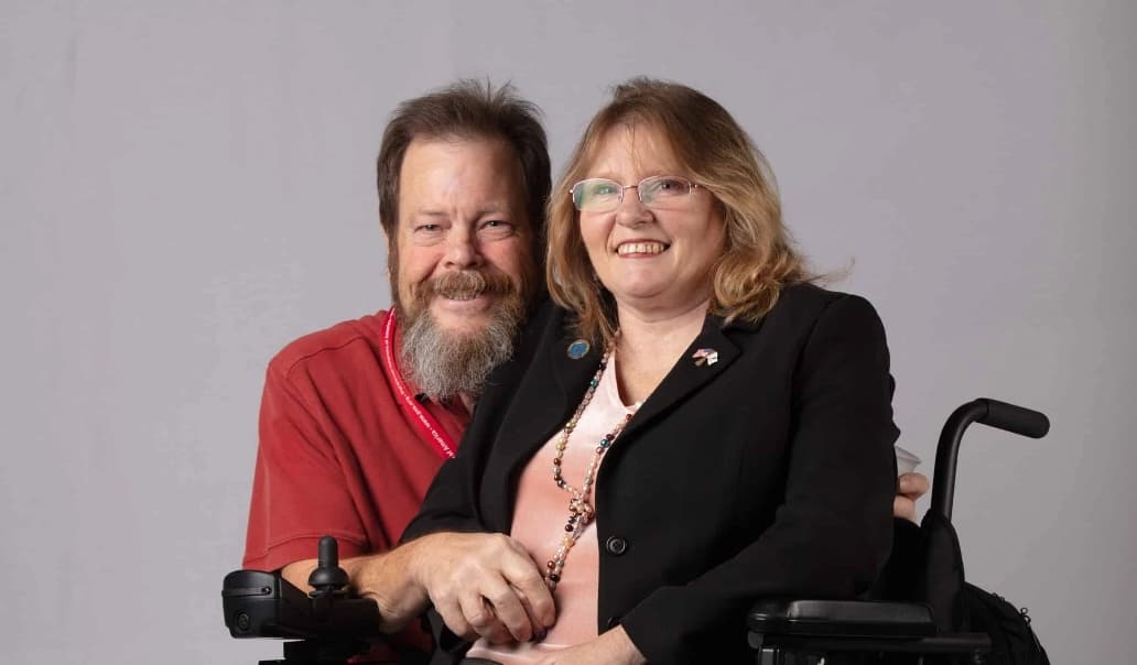 A smiling man with a beard and red shirt sits beside a smiling woman wearing glasses and a black blazer seated in a wheelchair. They are posing together against a plain light gray background.