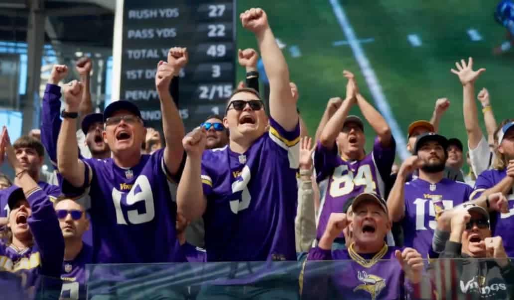 A group of excited Minnesota Vikings fans, dressed in purple jerseys, cheer passionately in the stands of a football stadium with a scoreboard and field visible in the background.