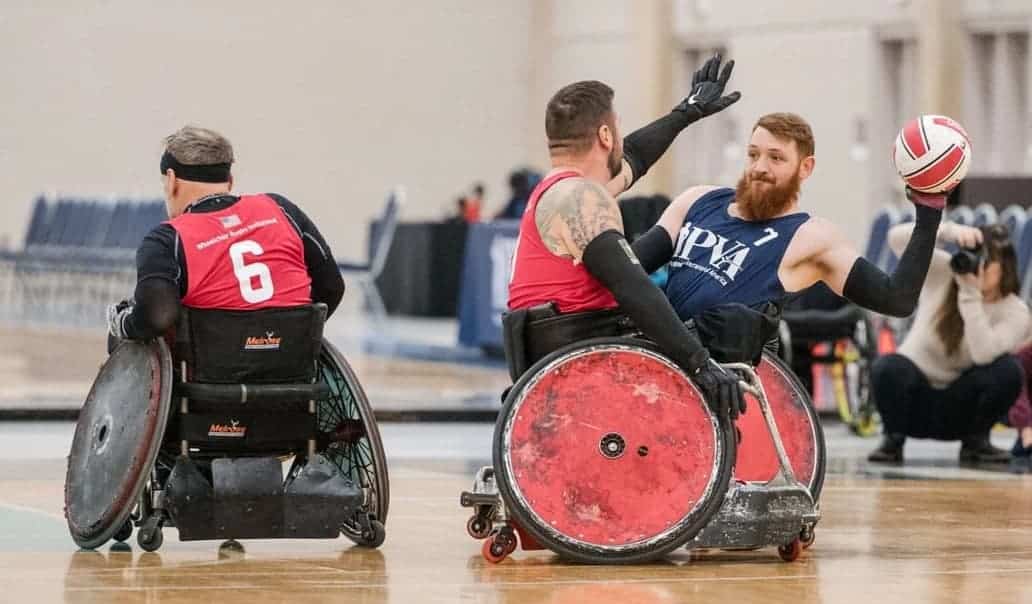 Two athletes in wheelchairs compete in a rugby match. One player in red blocks while another in blue prepares to throw the ball. A photographer kneels nearby capturing the action.