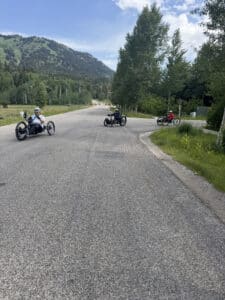 Three people ride recumbent tricycles and a bicycle on a wide paved road surrounded by green grass, trees, and distant hills under a partly cloudy sky.