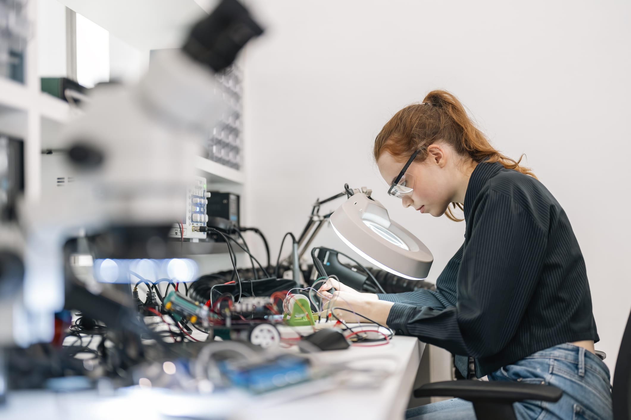 A woman wearing safety glasses works with electronic components at a lab bench, using tools and surrounded by equipment like a microscope and a magnifying lamp.