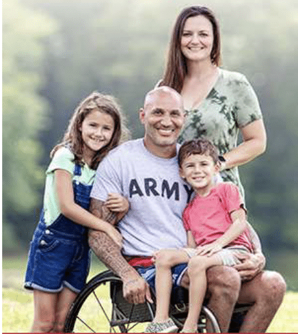A smiling man in a wheelchair wearing an "Army" shirt poses outdoors with a woman standing behind him and two young children beside him, all looking happy together.