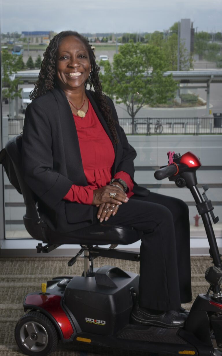 A woman wearing a black suit and red blouse smiles while sitting on a red mobility scooter indoors, with large windows showing a view of trees and buildings outside.