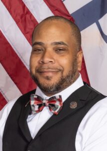 Marcus Murray smiles while wearing a checkered bow tie, white shirt, and black vest with pins. An American flag and another flag are visible in the background.