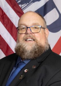 Matthew Peeling in a dark suit and a blue shirt smiles in front of a large American flag background.