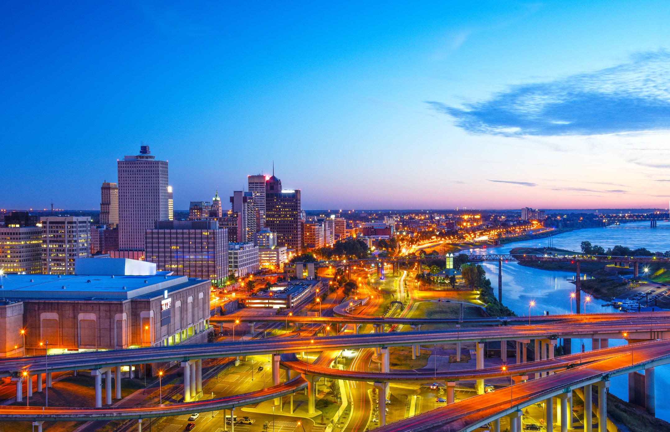 A vibrant cityscape of Memphis at dusk, with illuminated skyscrapers, highways, and bridges curving over the Mississippi River under a blue and orange sky.