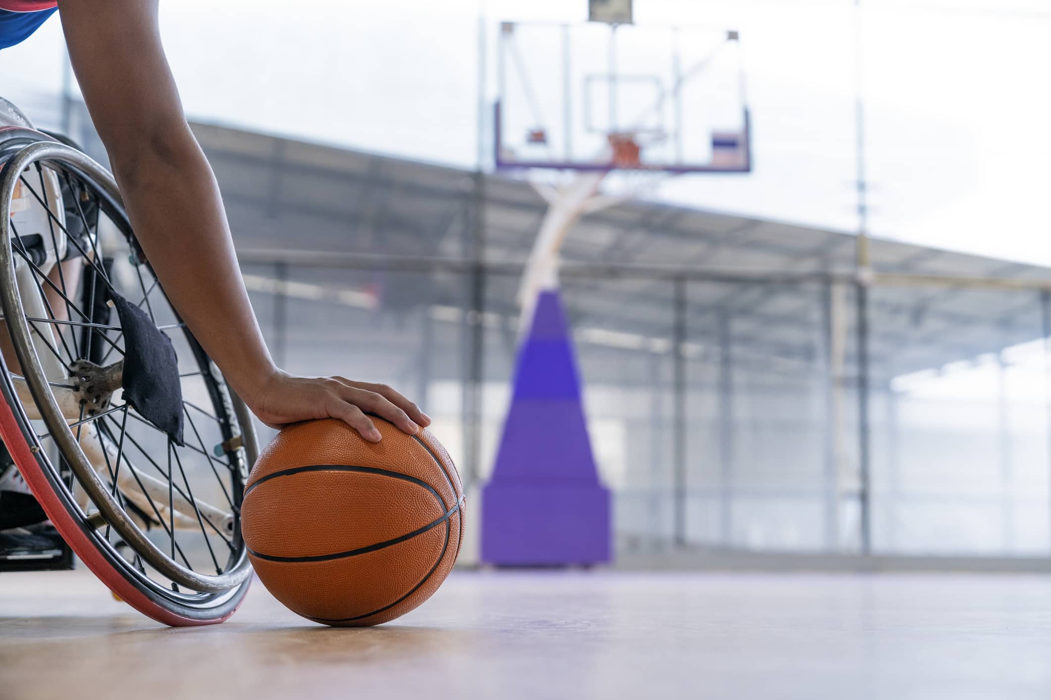 A person in a wheelchair prepares to dribble a basketball on an indoor court, with a hoop and backboard visible in the background.