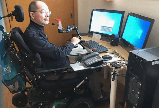 A man in a motorized wheelchair uses a computer setup with two monitors, a keyboard, and adaptive technology, including a mouth-operated device, in a home office environment.