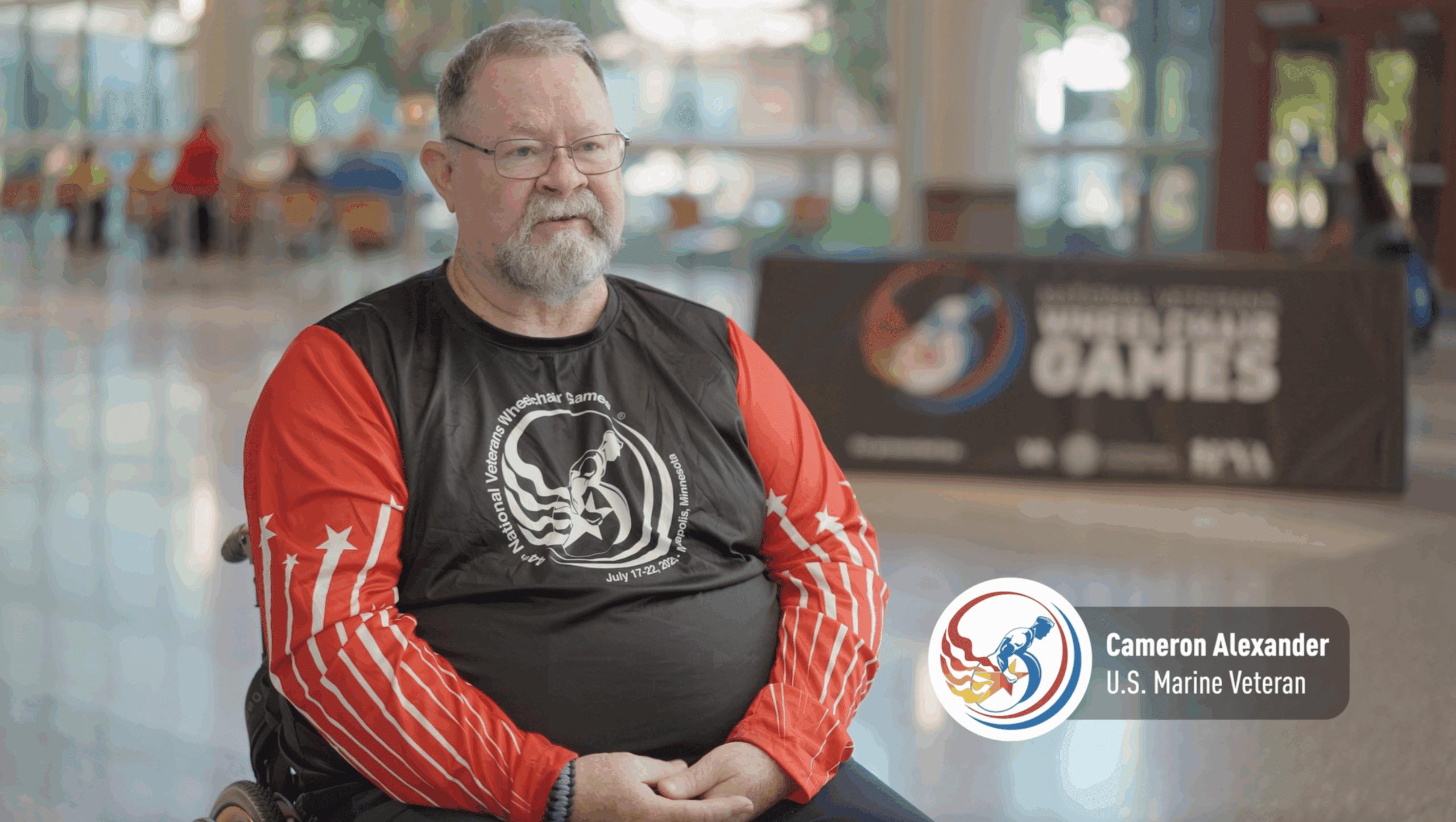 A man in a wheelchair wearing a red and black sports shirt sits indoors, with a blurred banner for the National Veterans Wheelchair Games in the background. A label reads: "Cameron Alexander, U.S. Marine Veteran.