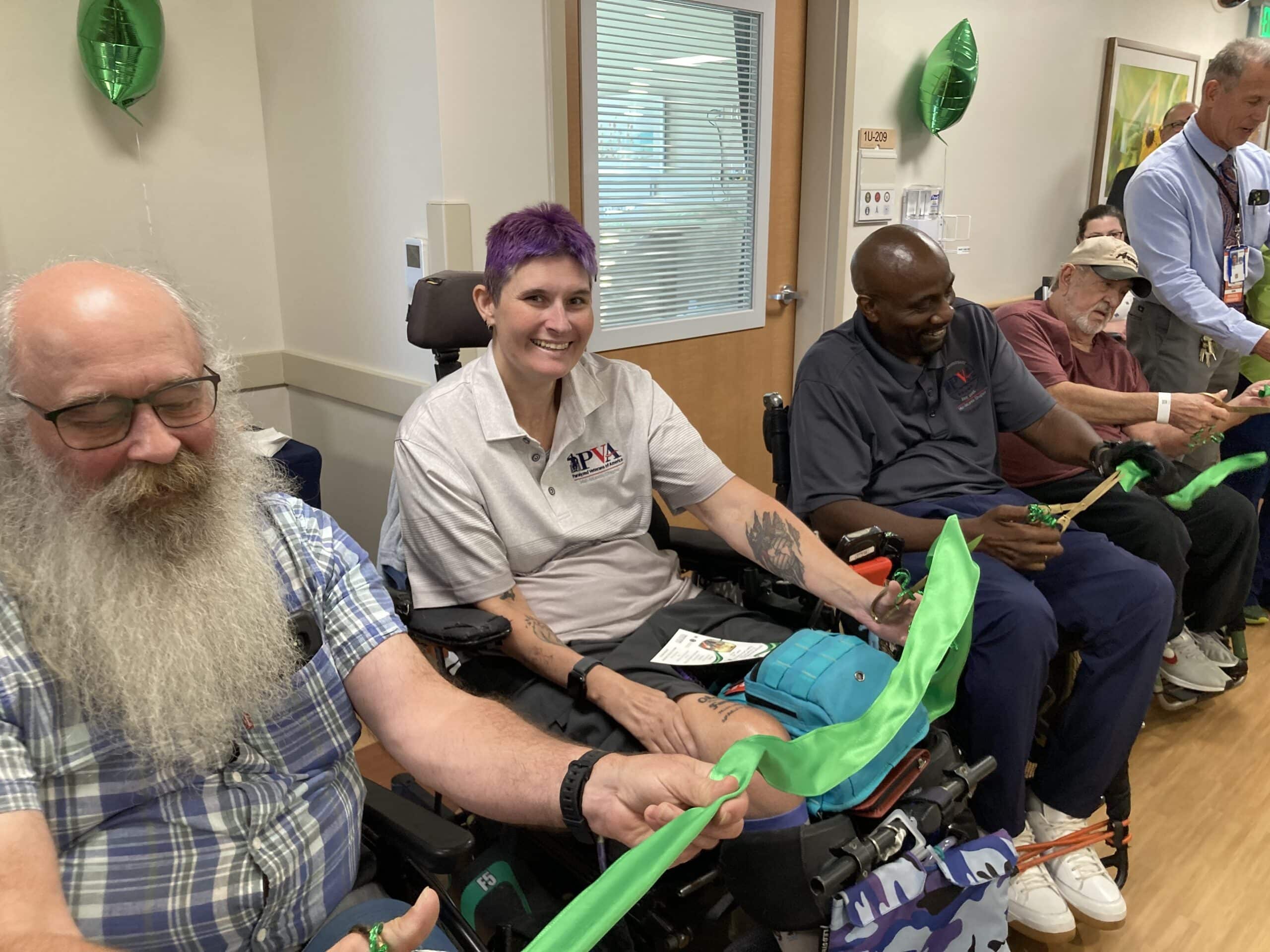 Several people, some in wheelchairs, smile and hold a long green ribbon at an indoor event. Green balloons and staff are visible in the background, creating a celebratory atmosphere.