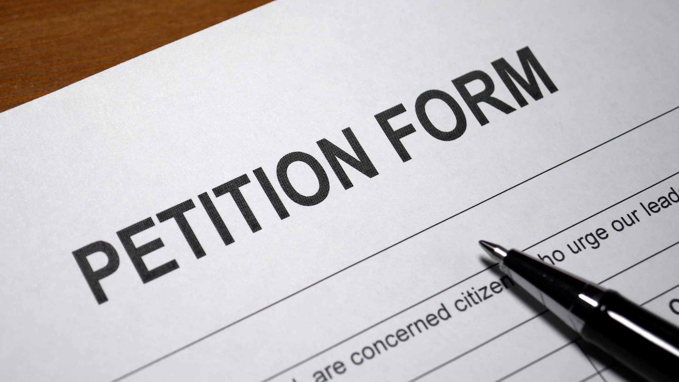 A close-up of a petition form on a wooden surface, with the words "PETITION FORM" in bold at the top and a black pen resting on the paper.