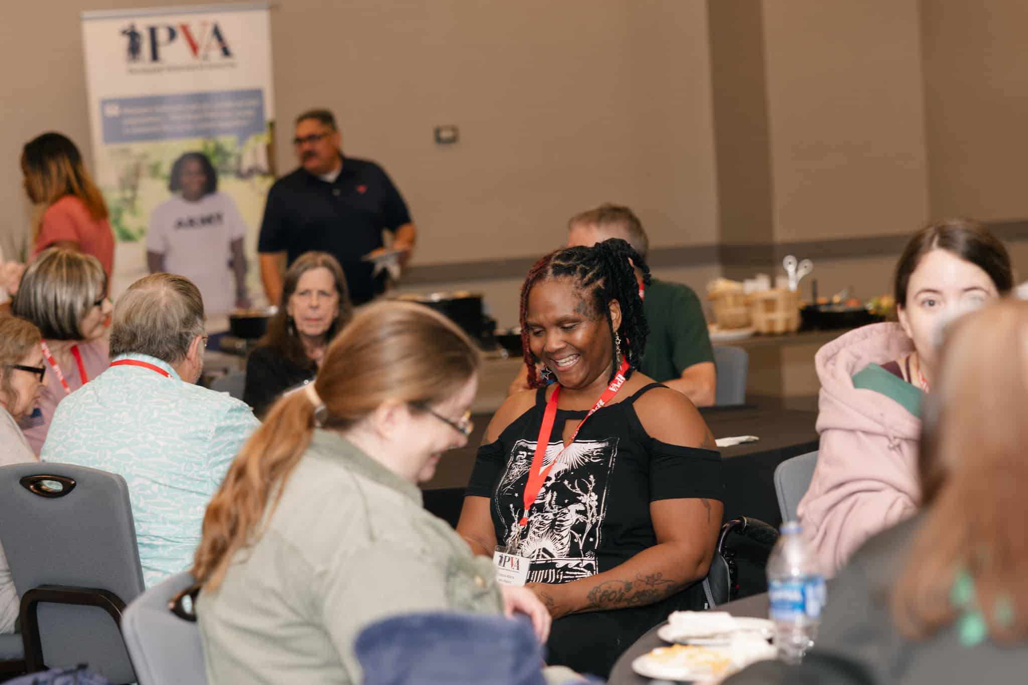 A group of female Veterans sit around tables at an indoor event, talking and smiling. Food and drinks are on the tables. A banner for "PVA" is visible in the background. The atmosphere appears friendly and social.