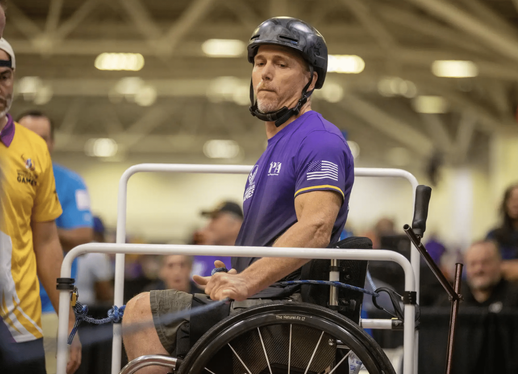 A man wearing a helmet and a purple shirt sits in a wheelchair, participating in a competitive event indoors, with spectators and other participants visible in the background.