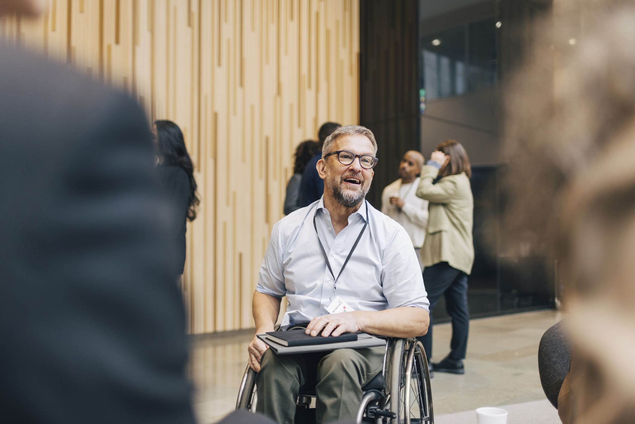 A man in a wheelchair, wearing glasses and a lanyard, smiles while holding a tablet. He is in a modern indoor setting with people chatting and mingling in the background.