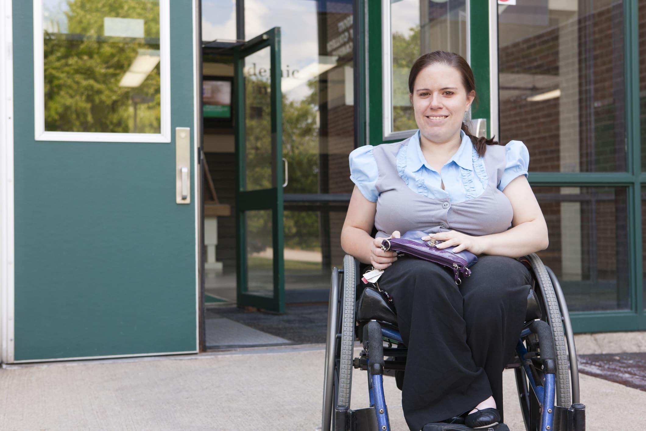 A woman in a wheelchair smiles while sitting outside a building with glass doors and brick walls. She holds a small purple bag and is dressed in a blue blouse and black pants.