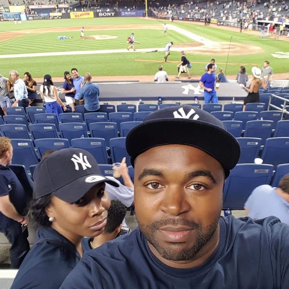 A man and woman wearing New York Yankees hats take a selfie in a stadium with a baseball field in the background, surrounded by other fans and empty seats.