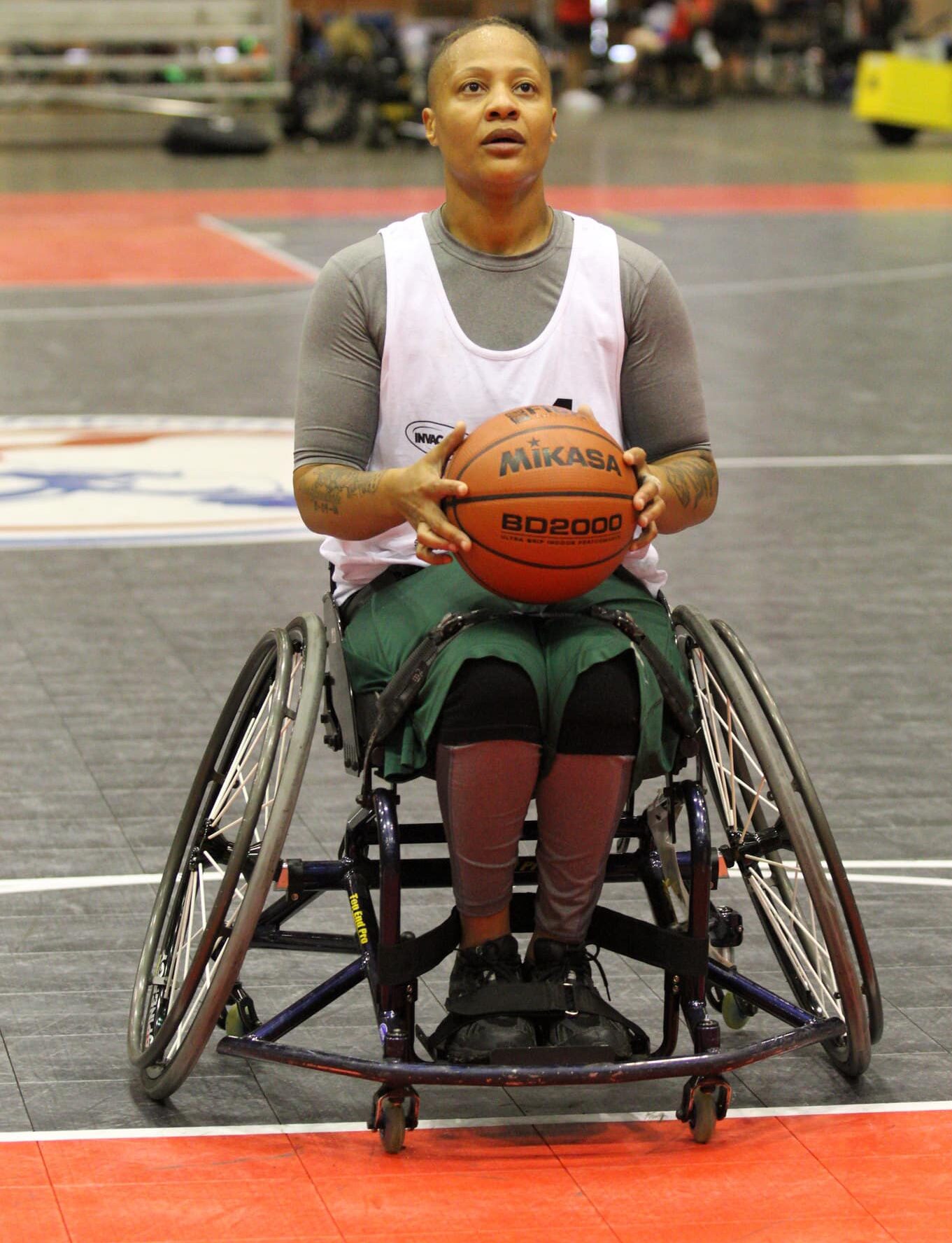 A basketball player in a wheelchair holds a basketball in both hands, preparing to take a shot on an indoor court. The player wears a white jersey and green shorts.